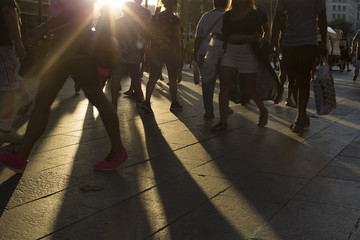 Crowds walking in a busy city district as the sun flares between them in the late afternoon creating long shadows on the ground
