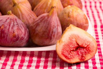 Group of figs in a white plate, with one cut fig on red tablecloth 
