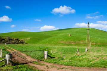 Fototapeta na wymiar Farmland with green hills, gate on dirt track and old windmill