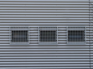 Three barred windows on a grey facade made of corrugated iron.