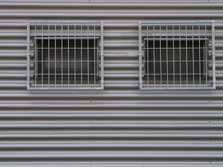 Two barred windows on a grey facade made of corrugated iron.