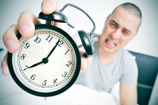 angry young man in bed holds his alarm clock