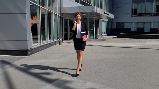Young business woman walking to the office early in the morning and talking by smartphone