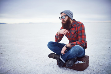 pensive contemplative hipster stroking awesome manly beard sitting on retro suitcase in salt flats all alone