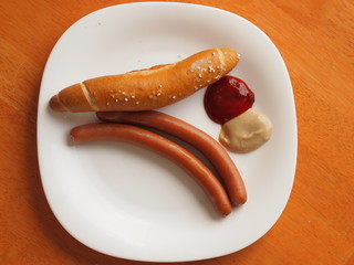 Two wiener sausages with a white bread roll, ketchup and mustard on a white plate that is placed on a wooden table.