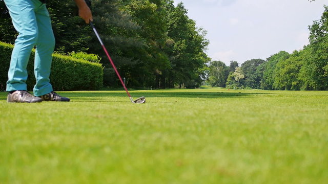 A man dressed with a blue trouser is playing a drive at a golf course on a summer day. The camera is placed on the grass and turning slowly from left to right. 