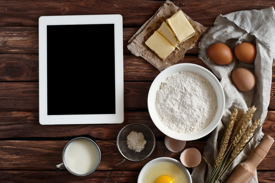 White tablet computer with blank screen with ingredients for making pancakes or cake - flour, egg, butter, milk on the wooden background. top view. rustic style. free space for text . Copy space