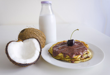 Breakfast and brunch coconut pancakes meal with cracked open coco, glass bottle of milk and cherry on white background. Stack of crepes is topped with cream syrup ready to eat.