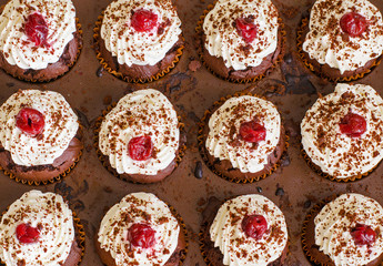 top view of many freshly baked chocolate muffins with whipped cream and cherries on top in a baking form