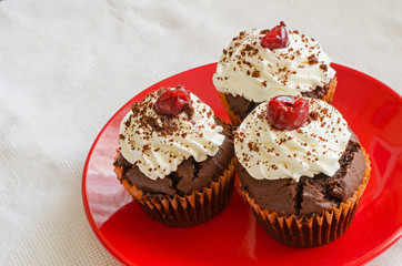 three chocolate muffins with whipped cream and cherry on top on a red plate on a white table cloth 
