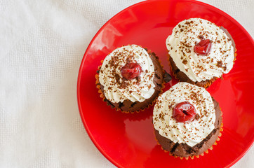 top view of three chocolate muffins with whipped cream and cherry on top on a red plate on a white table cloth 