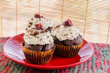 three chocolate muffins with whipped cream and cherries on a red plate in rustic environment