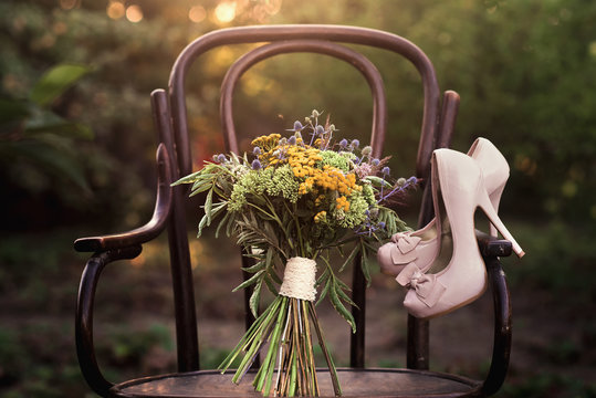 Beautiful wedding shoes with high heels and a bouquet of colorful flowers on a vintage chair on the nature in sunset light, decorations, preparing for the wedding, details, boudoir