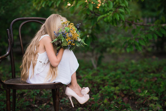 The little girl in high-heeled shoes large size in a white dress standing on a chair with a vintage bouquet in the hands of fashio