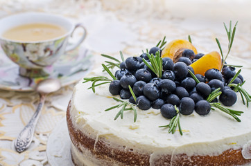 side view of delicious home made layered cake with cream, berries, rosemary and nectarine on top on a decorative white table cloth next to a vintage cup of tea
