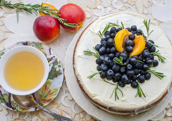 top view of delicious home made cake with cream, berries, rosemary and nectarine on top on a decorative white table cloth next to a vintage cup of green tea and nectarines
