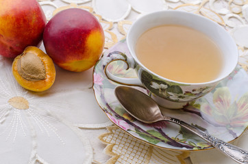 green tea in a vintage decorative cup with metal spoon and fitting plate next to peaches and nectarines on a white ornamental table cloth