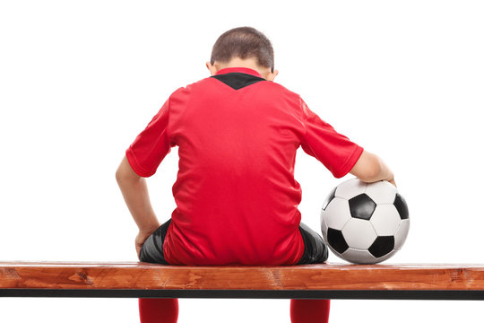 Sad little boy in red soccer jersey seated on a bench and holdin