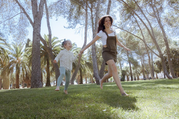 Naklejka na ściany i meble Una niña y su mama corriendo por el parque. Madre e hija jugando en el parque. Una niña y su mama disfrutando del verano rodeadas de árboles. Madre joven soltera.