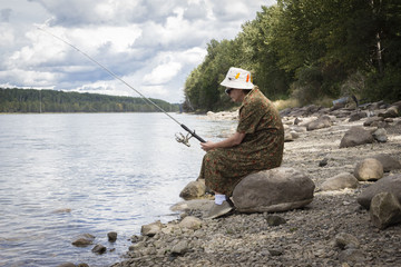 horizontal image of a middle aged woman wearing  a fishing hat sitting on the banks of the river on a big stone waiting for the fish to bite on a hot summer day.