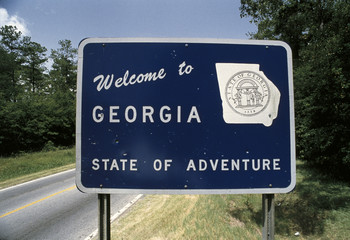 This is a road sign that says, welcome to Georgia, state of adventure. There is a white map of the...