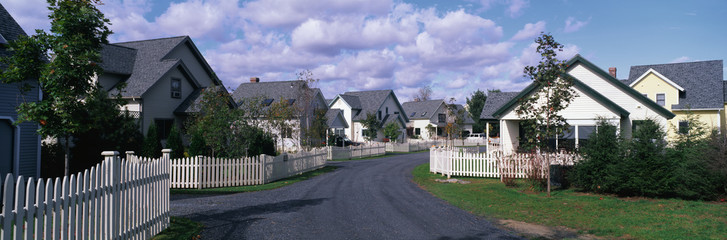 Fototapeta na wymiar This is a typical suburban American neighborhood. There are single family homes with a white picket fences in front of each house. A road leads down the center of the image that takes you past each house. There are trees beside each house.