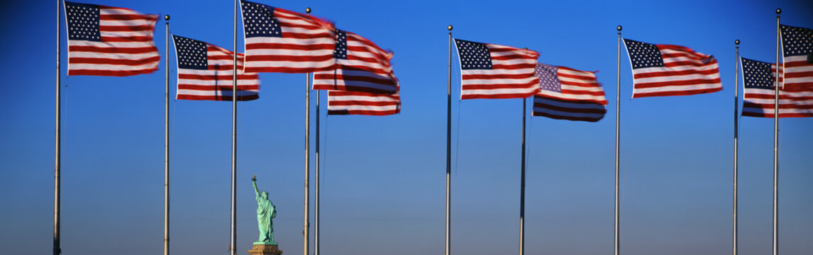 This is an image of the Statue of Liberty situated in between a group of American flags on flagpoles in Liberty Park, New Jersey. The flags are waving in the wind against a background of blue sky in the afternoon.
