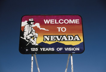 This is a road sign that says, Welcome to Nevada, 125 years of Vision. It is located on Interstate 15. It shows a miner with a pick in his hand, squatting next to the mountains against a red, orange and blue background. There is also a cloud in the sky.