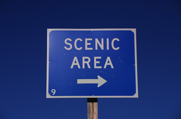 This is a road sign that says, Scenic Area, with a white arrow pointing to the right. The sign has a blue background and is against a blue sky.