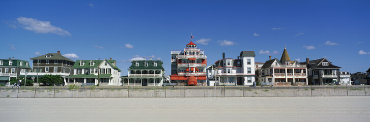These are Victorian style homes overlooking the beach in Cape May. There is wooden fence separating the beach and the houses. The homes have large front porches. The sky is a deep blue with just a few white puffy clouds.