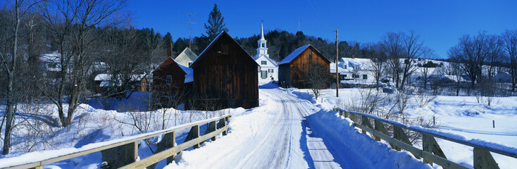 This is a small town in New England showing a white Methodist Church with steeple. There is a snow covered bridge in the foreground with older looking brown wood buildings behind it. The bare trees of winter surround the buildings. - Powered by Adobe