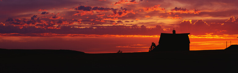 This is a farm at sunset. The farm house is silhouetted against an orange sky. - obrazy, fototapety, plakaty