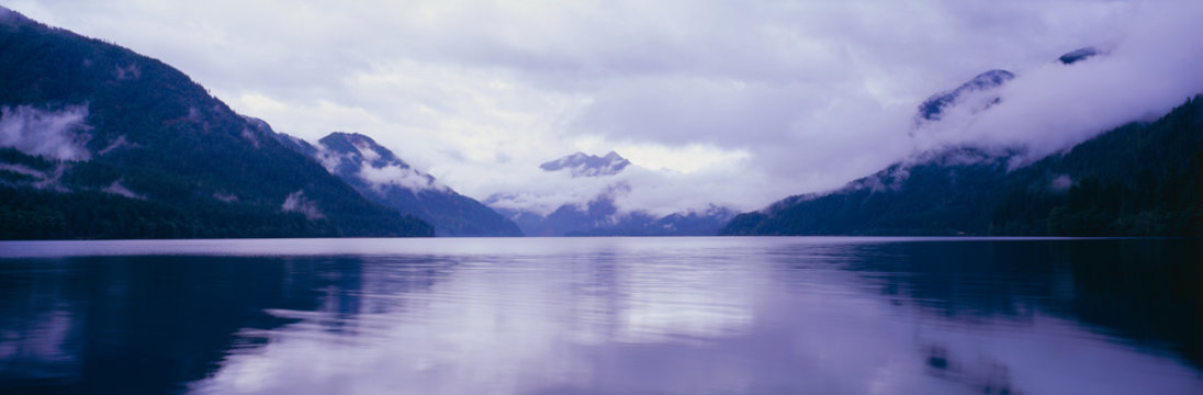 This is an image of the Olympic Peninsula with Crescent Lake in the foreground. The mountains in the background are covered with low altitude clouds. There is a reflection in the lake of the clouds and mountains. The sky is overcast with clouds.