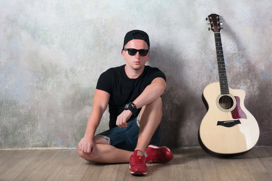 Man in denim shorts sitting next to a guitar on the wall background in style grunge, music, musician, hobby, lifestyle, hobby