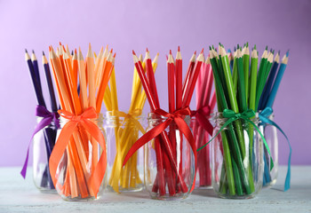Bright pencils in glass jars on wooden table, on purple background
