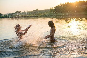 Two happy slim young girls splashing in lake at sunset