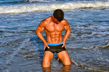 Fototapeta premium Strong bodybuilder with six pack.Fitness trainer with perfect abs, shoulders,biceps, triceps,chest, flexing his muscles on the beach with sea waves on the background, training in vacation