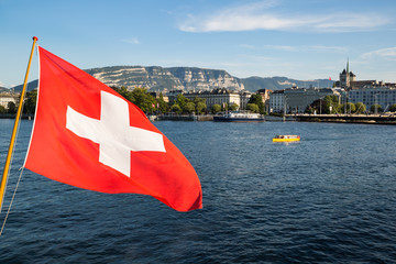 A Swiss national flag floating in the wind over the Lake Geneva, with the city of Geneva in the background