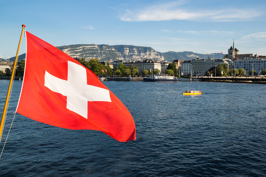 A Swiss National Flag Floating In The Wind Over The Lake Geneva, With The City Of Geneva In The Background