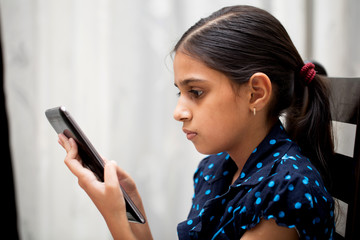ten year girl playing with her tablet pc or tab in her house sitting on a sofa or a wooden chair...