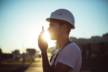 Engineer builder using a walkie talkie giving instructions at a construction site. Sunset time.