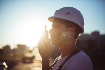 Engineer builder using a walkie talkie giving instructions at a construction site. Sunset time.