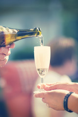 An image of someone pouring champagne to a glass in the summer and outdoors. Image has a vintage effect.