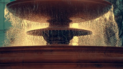 A slow-motion backlit scene of water dropping from a fountain. The intense sun background light creates the illusion of golden droplets of water, providing the whole scene a timeless aura.
