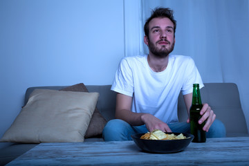 Young man watching TV at nighttime with chips and beer