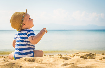 Baby boy with hat sitting on the beach. - 90129833