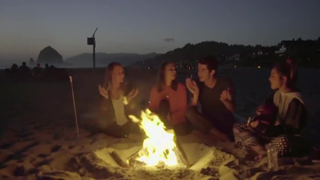Friends clap to music around a camp fire at the beach 