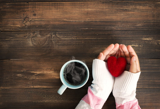 Above view of female hand holding red heart with hot cup of coffee on wood table. Photo in vintage color image style.
