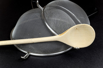 Two kitchen sieves with wooden spoon on black background