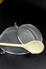 Two kitchen sieves with wooden spoon on black background